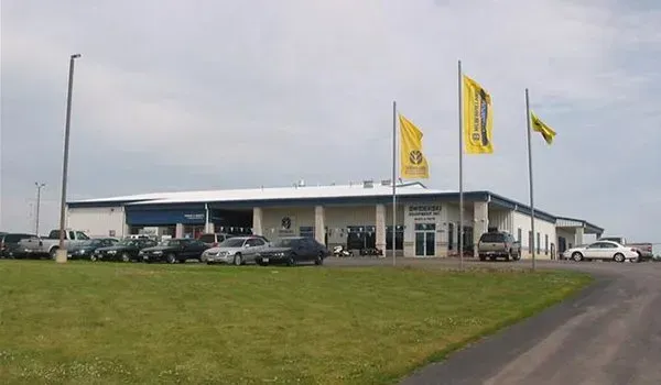 Low white commercial building with yellow flags, parked cars, and a grassy lot under cloudy skies