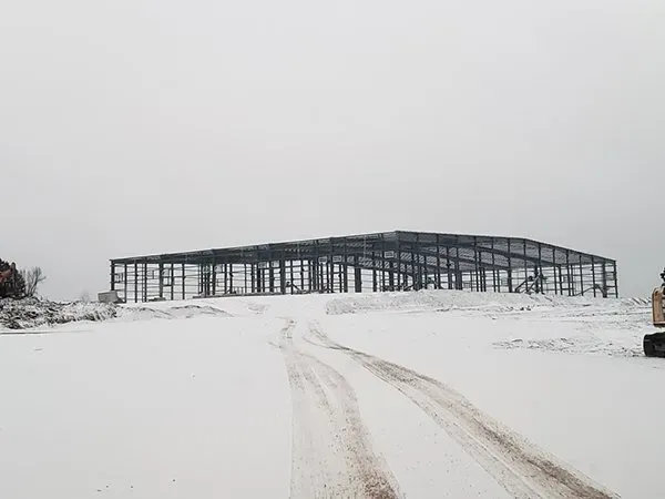 Snowy field with tire tracks leading to a large metal building under an overcast sky