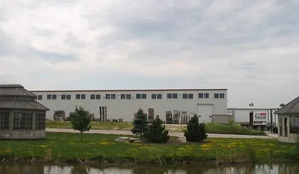 Wide industrial building beside a pond, with trees and grass in the foreground under an overcast sky.