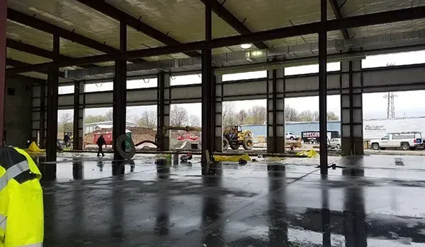 Partially built warehouse interior with workers, wet floor, and construction equipment near large open windows