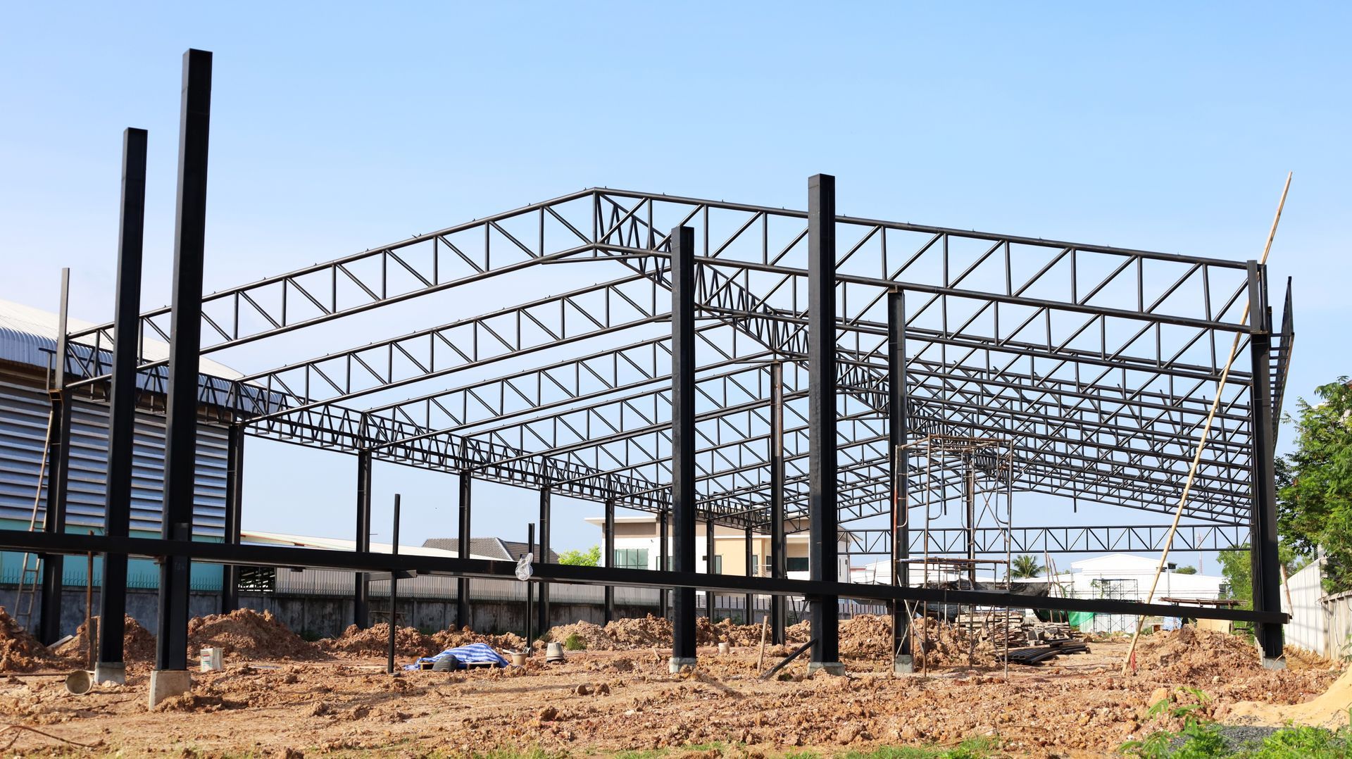 Steel frame of a large building under construction on a cleared site, with dirt and debris in front.