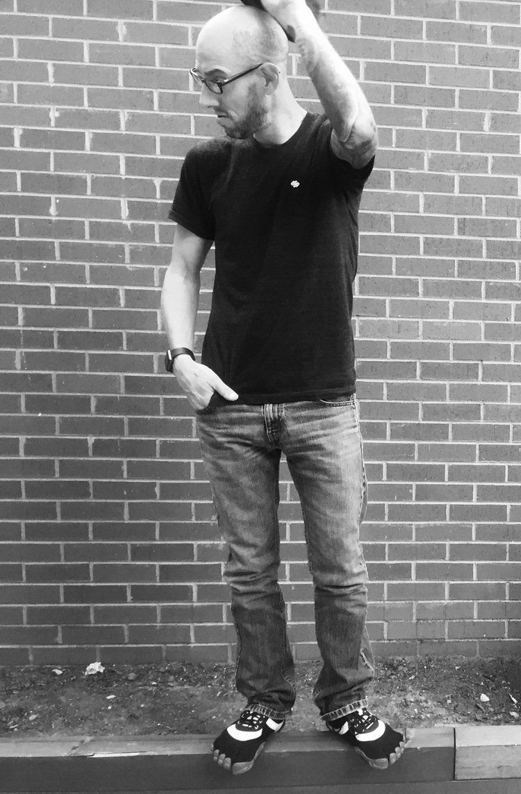 A man in a black shirt and jeans is standing in front of a brick wall.