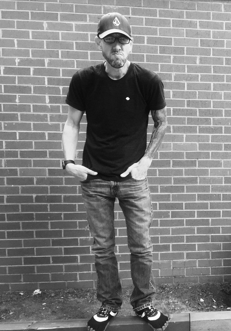 A man wearing a hat and a black shirt is standing in front of a brick wall.
