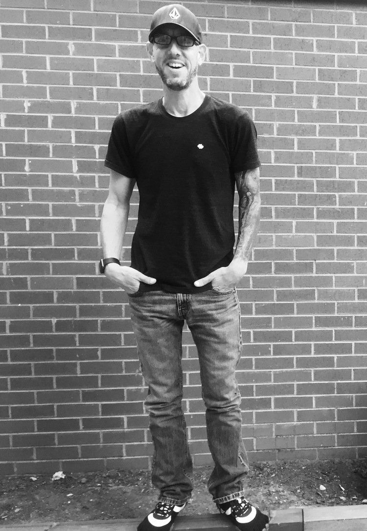 A man wearing a hat and sunglasses is standing in front of a brick wall.
