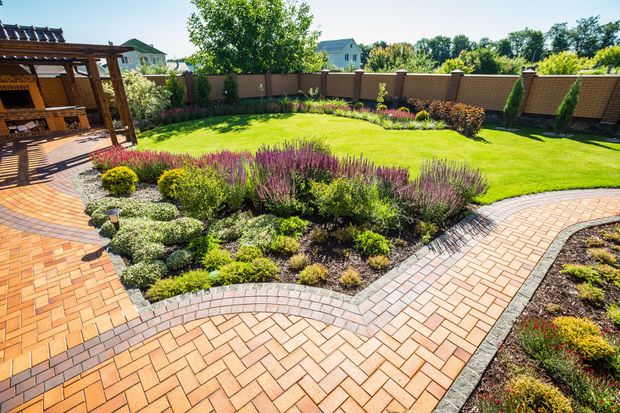 Curved brick path around a lush green backyard garden with colorful shrubs and a wooden pergola