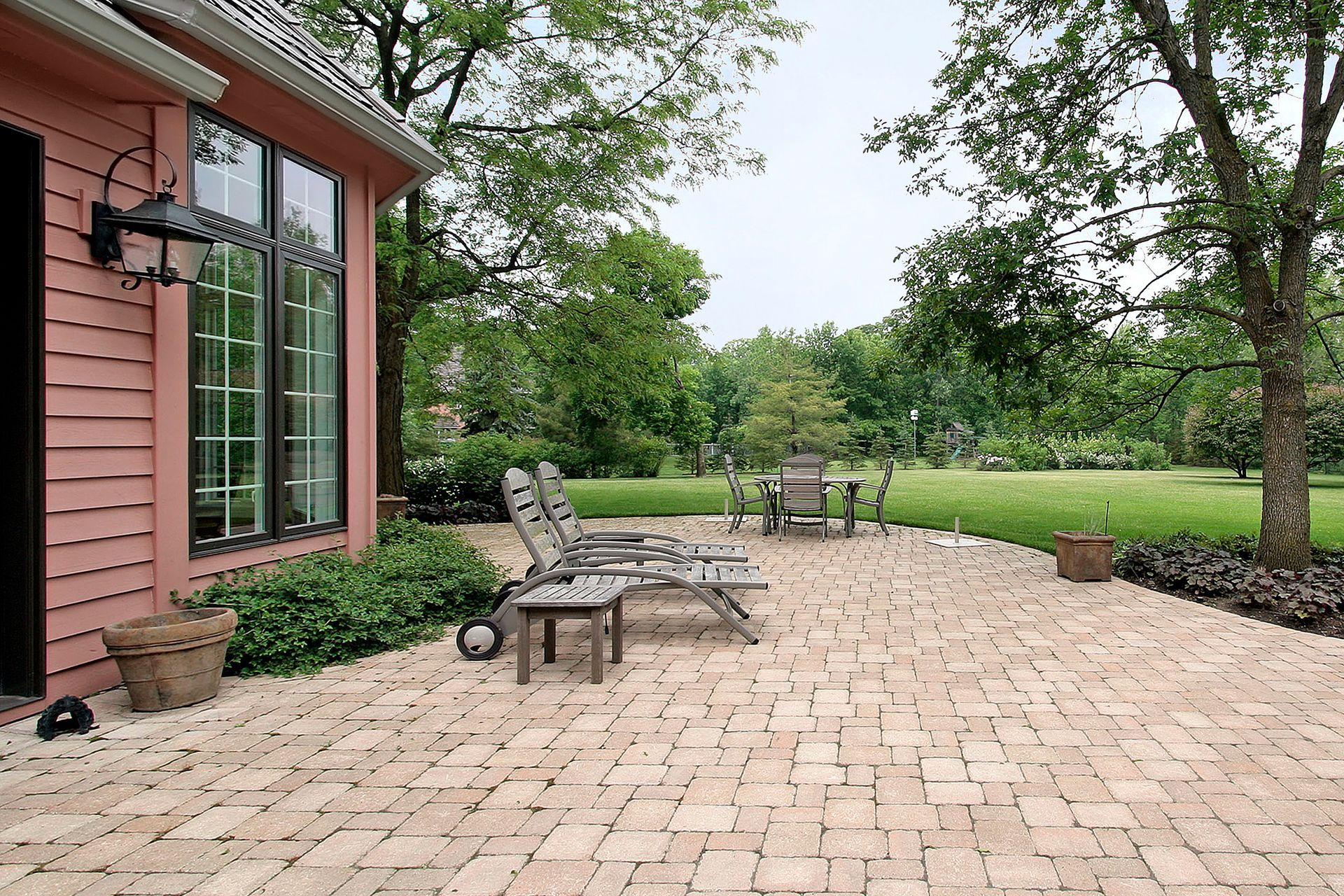Brick patio beside a red house with lounge chairs, potted plants, and a grassy yard under trees Brick patio beside a red house with lounge chairs, potted plants, and a grassy yard under trees