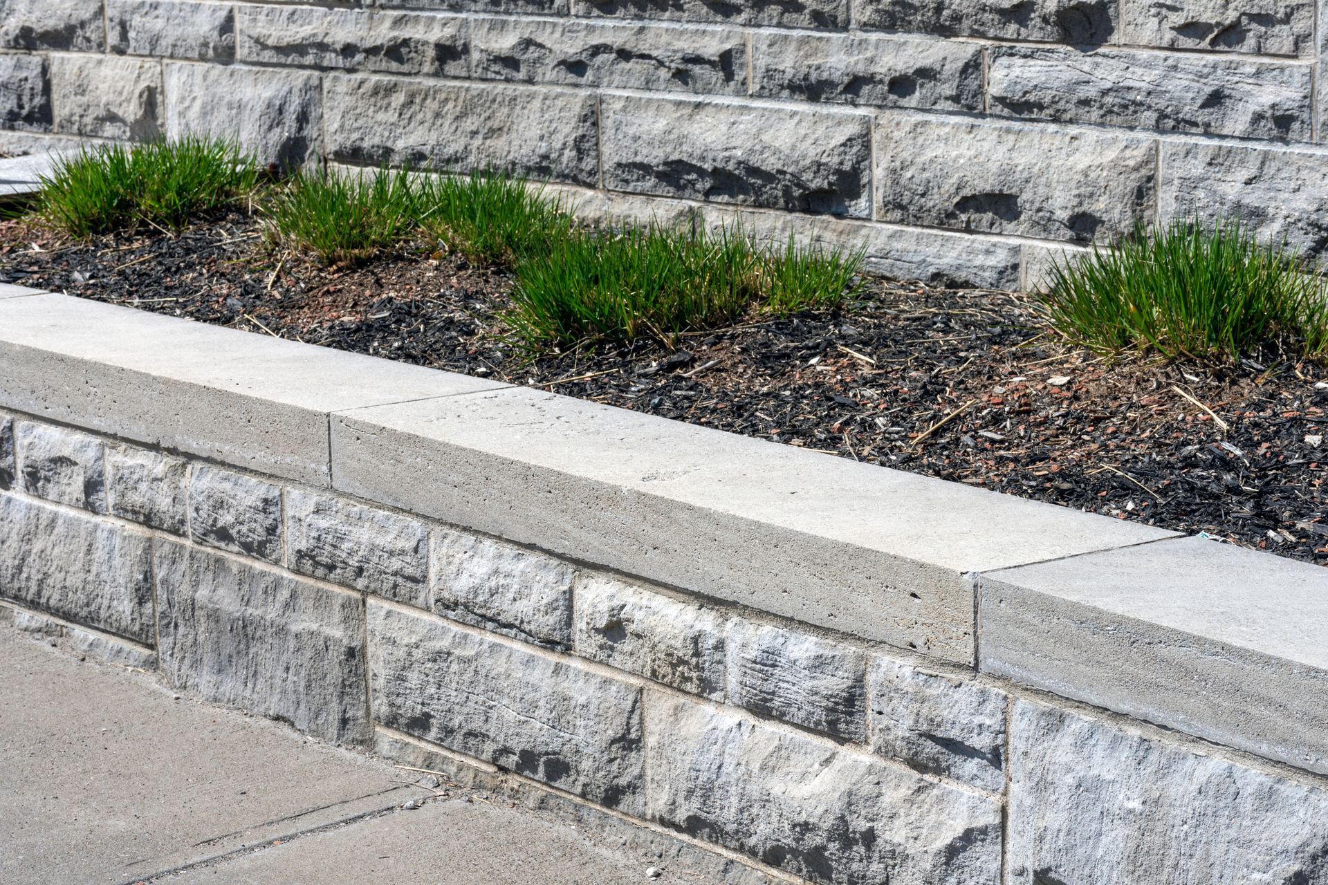 Stone retaining wall with stepped landscaping and small green plants in front of a paved walkway