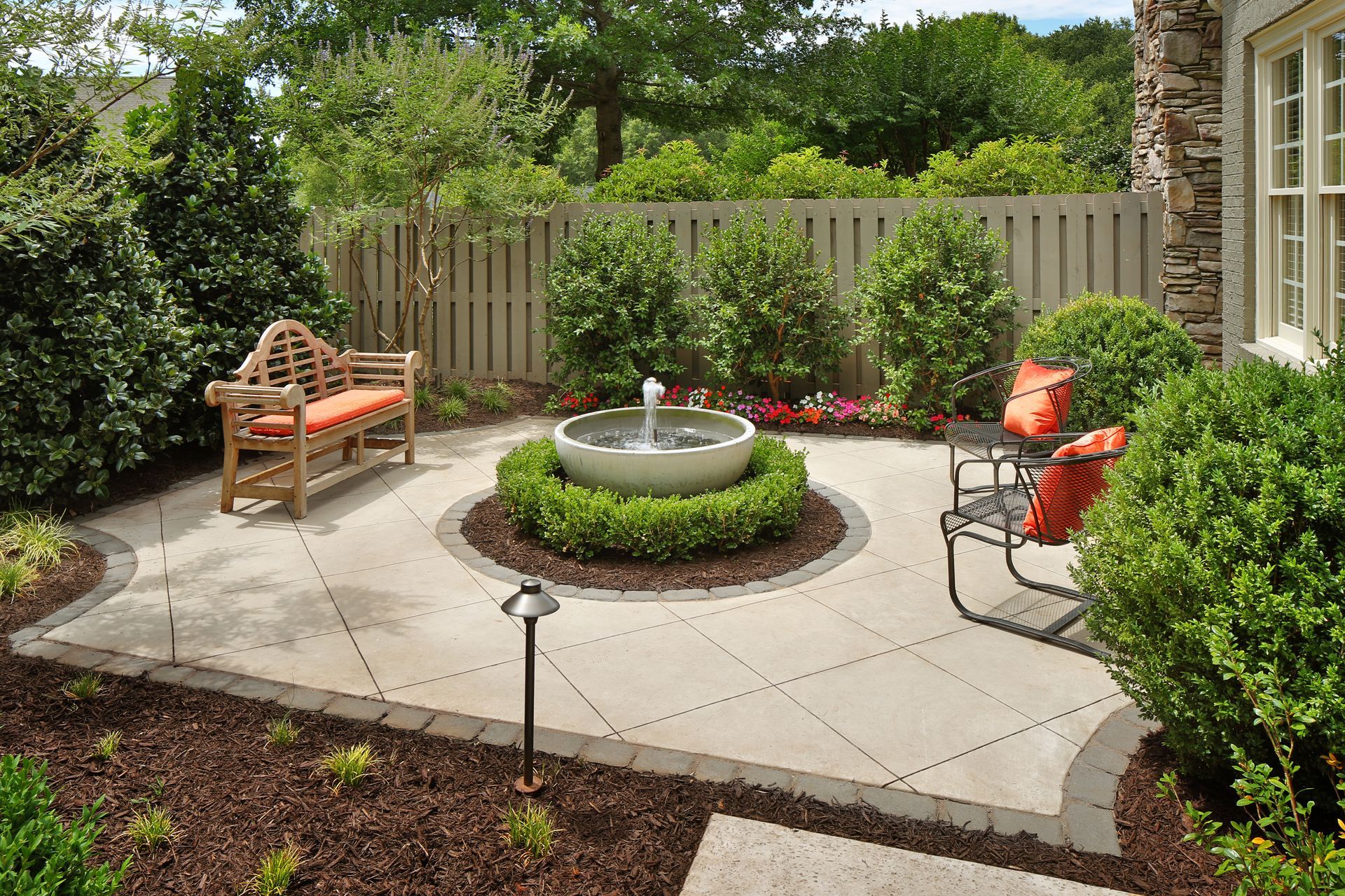 Small landscaped patio with a round fountain, bench, flower beds, and orange chairs beside a house Small landscaped patio with a round fountain, bench, flower beds, and orange chairs beside a house