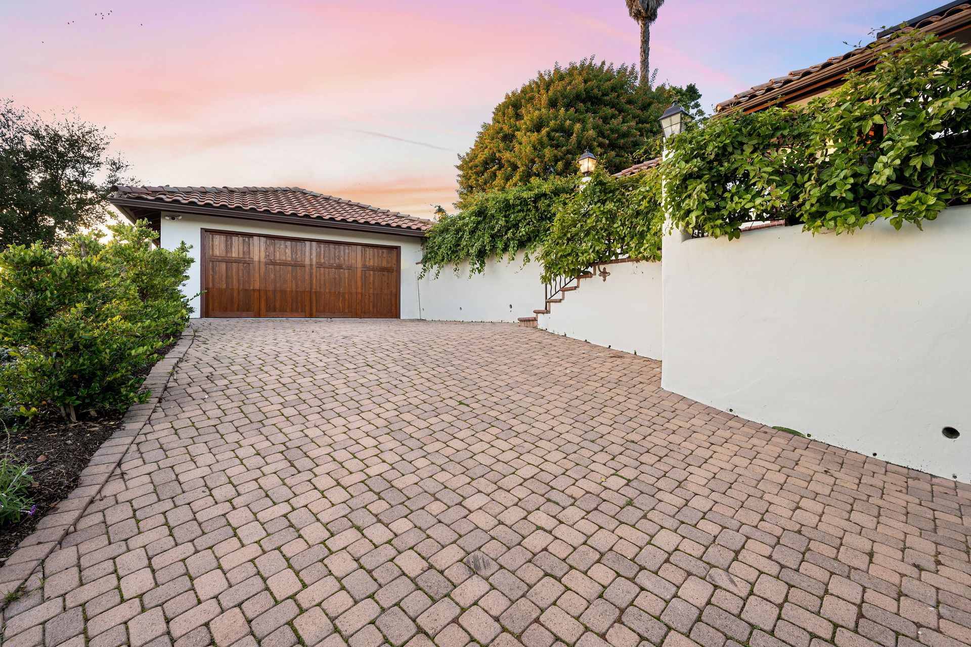 Paved driveway leading to a wooden garage door beside a white stucco wall and greenery at sunset.