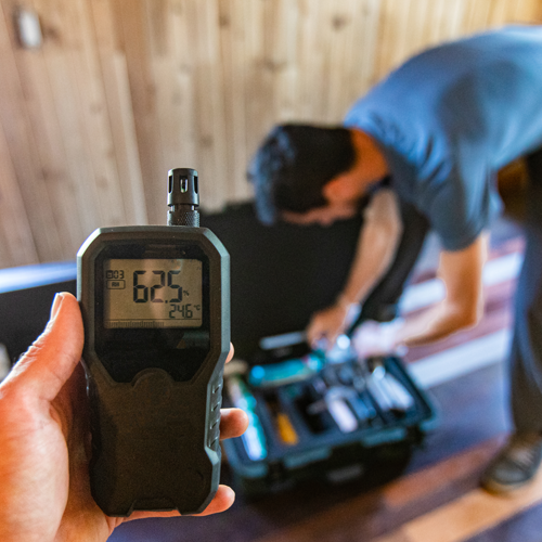 Handheld digital meter in foreground, with a person kneeling over tools in the background inside a wooden room