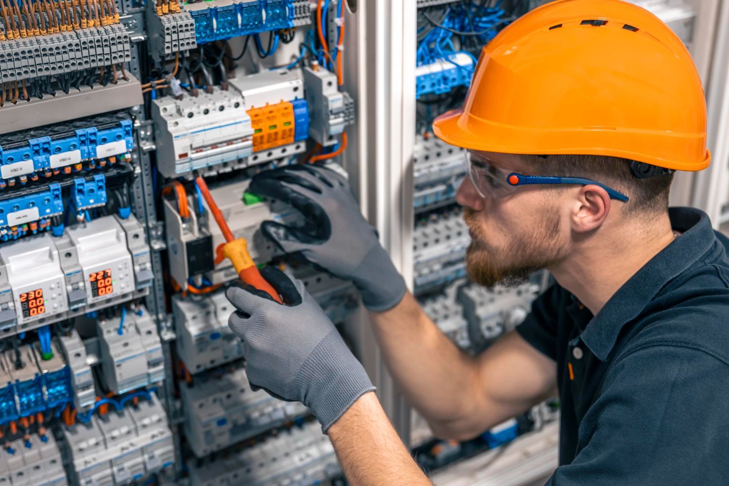 A man wearing a hard hat and gloves is working on an electrical panel.