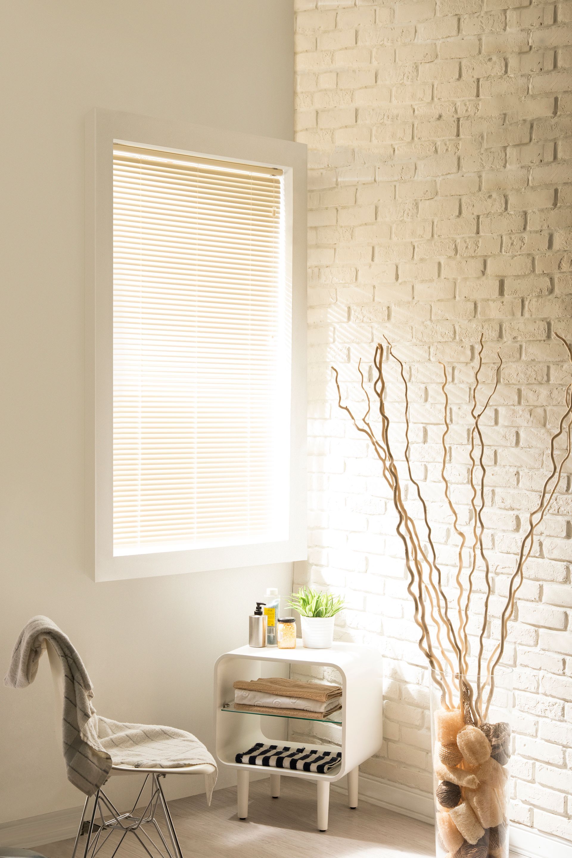Corner of a bright, modern room with white brick wall, window with blinds, and decorative twigs in a glass vase.