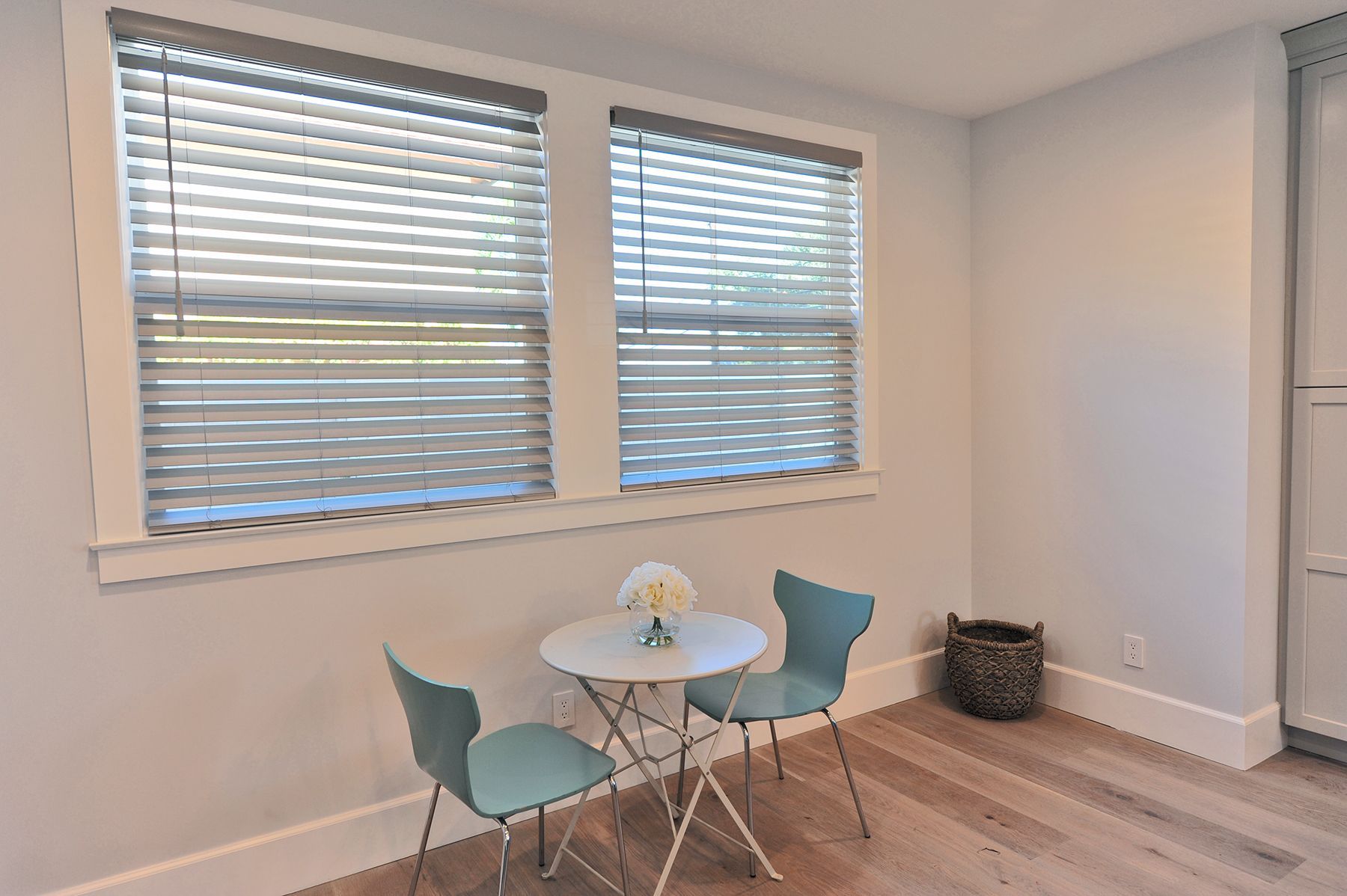 Small table and two teal chairs by a window with blinds, in a room with light wood floors.