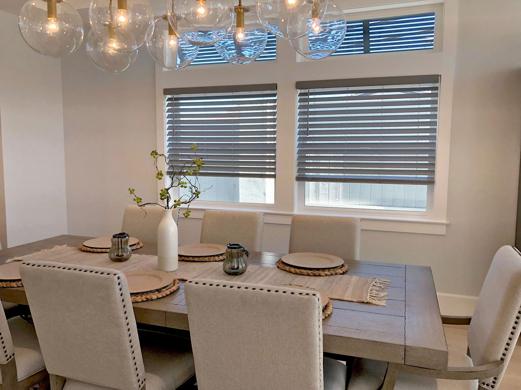 Dining room with a table set for a meal.  Gray blinds cover the windows. Chandelier above the table.