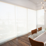 White roller shades cover windows in a dining room with a glass table and wooden chairs.