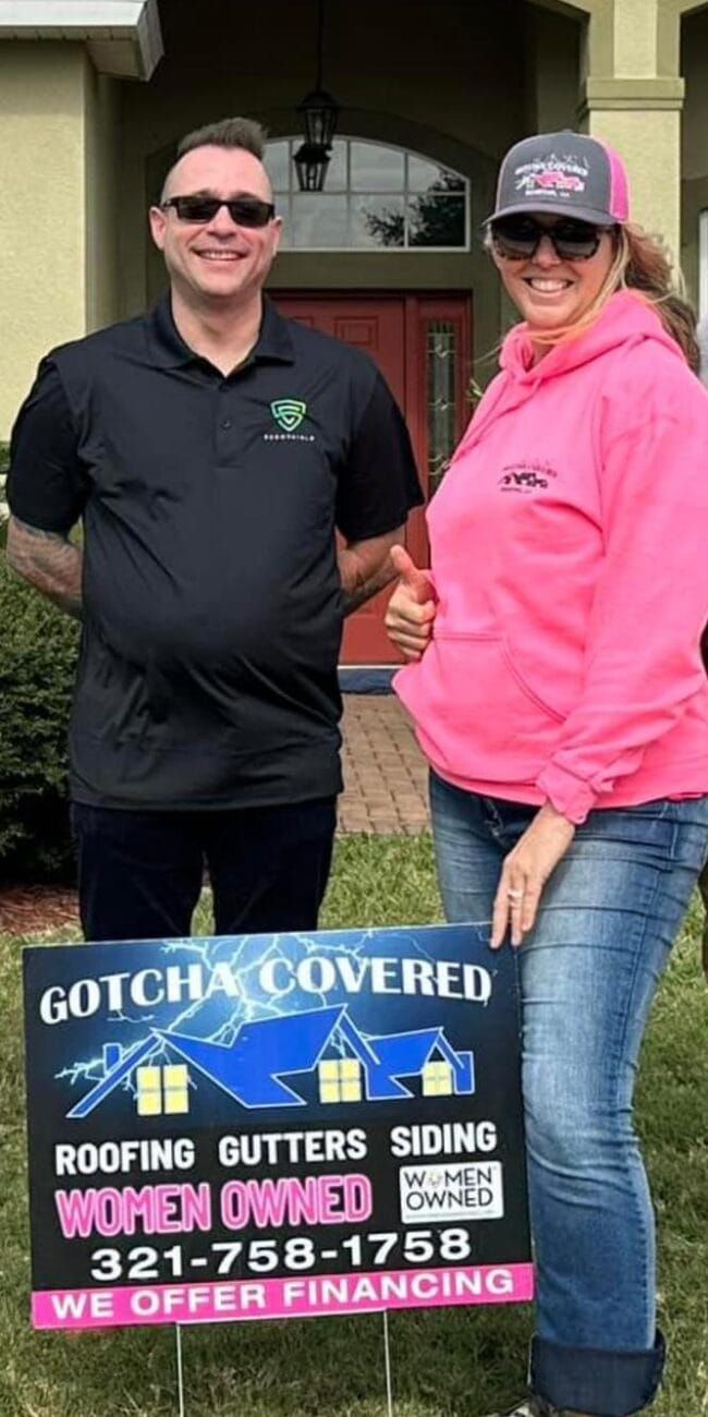 A man and a woman are standing in front of a house holding a sign.