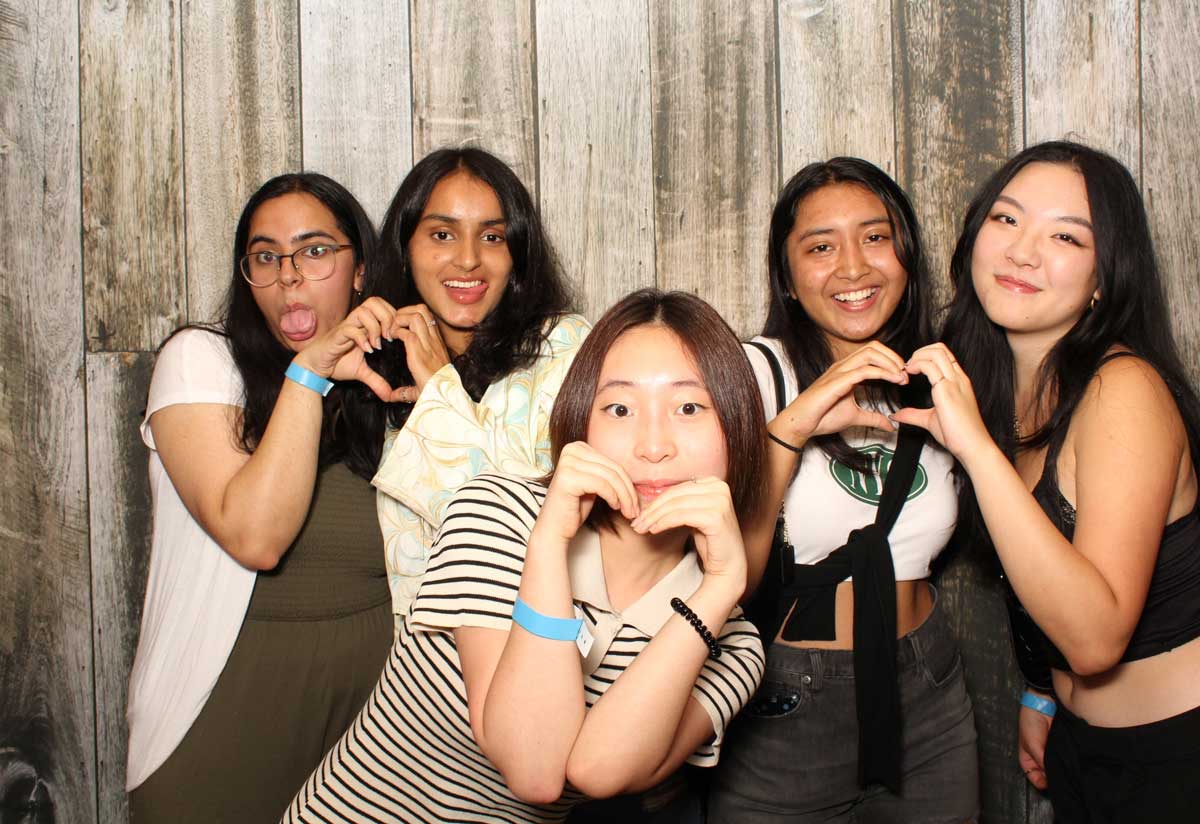 Five smiling people making heart shapes with their hands in front of a wood background.