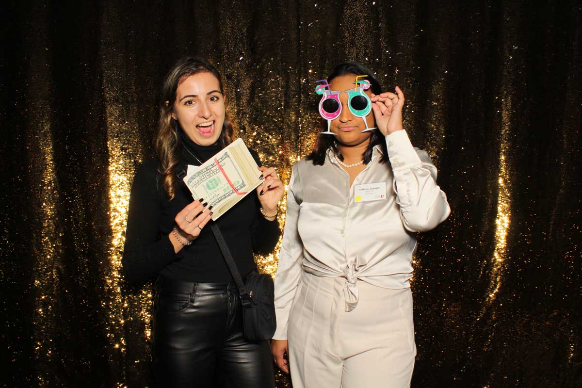 Two people posing in front of a gold sequin backdrop; one holds cash, the other wears silly glasses.