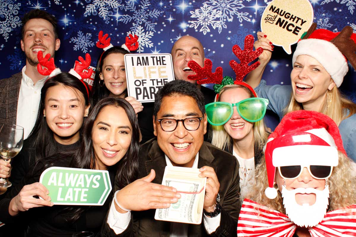 Group of people at a festive photo booth, holding props with humorous phrases, and wearing holiday accessories.