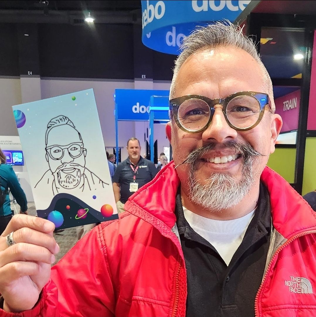 Man in glasses and red jacket holds drawing of himself; smiles. Indoors, convention backdrop.