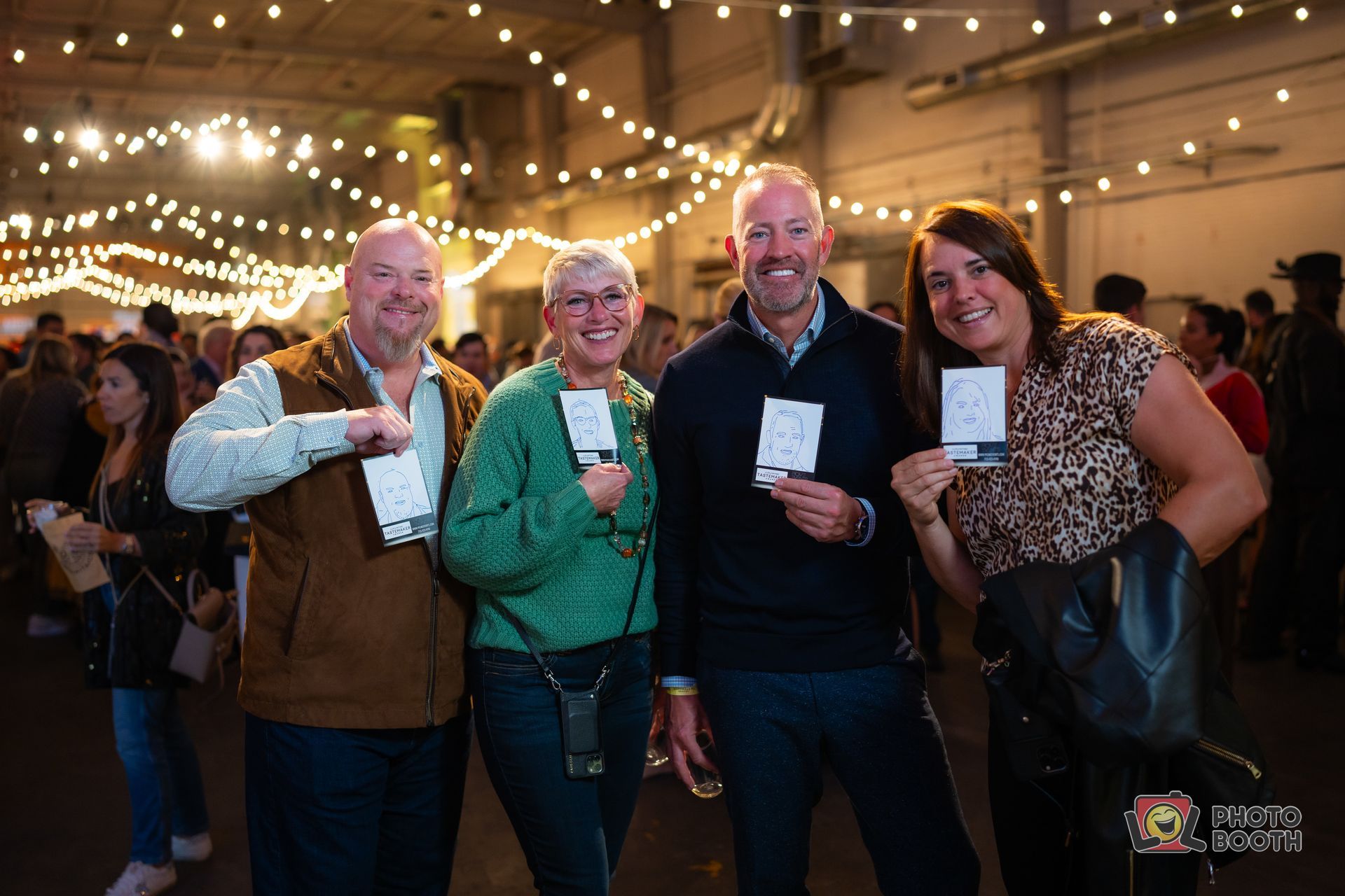 Four smiling people holding cards pose at an event lit with string lights.