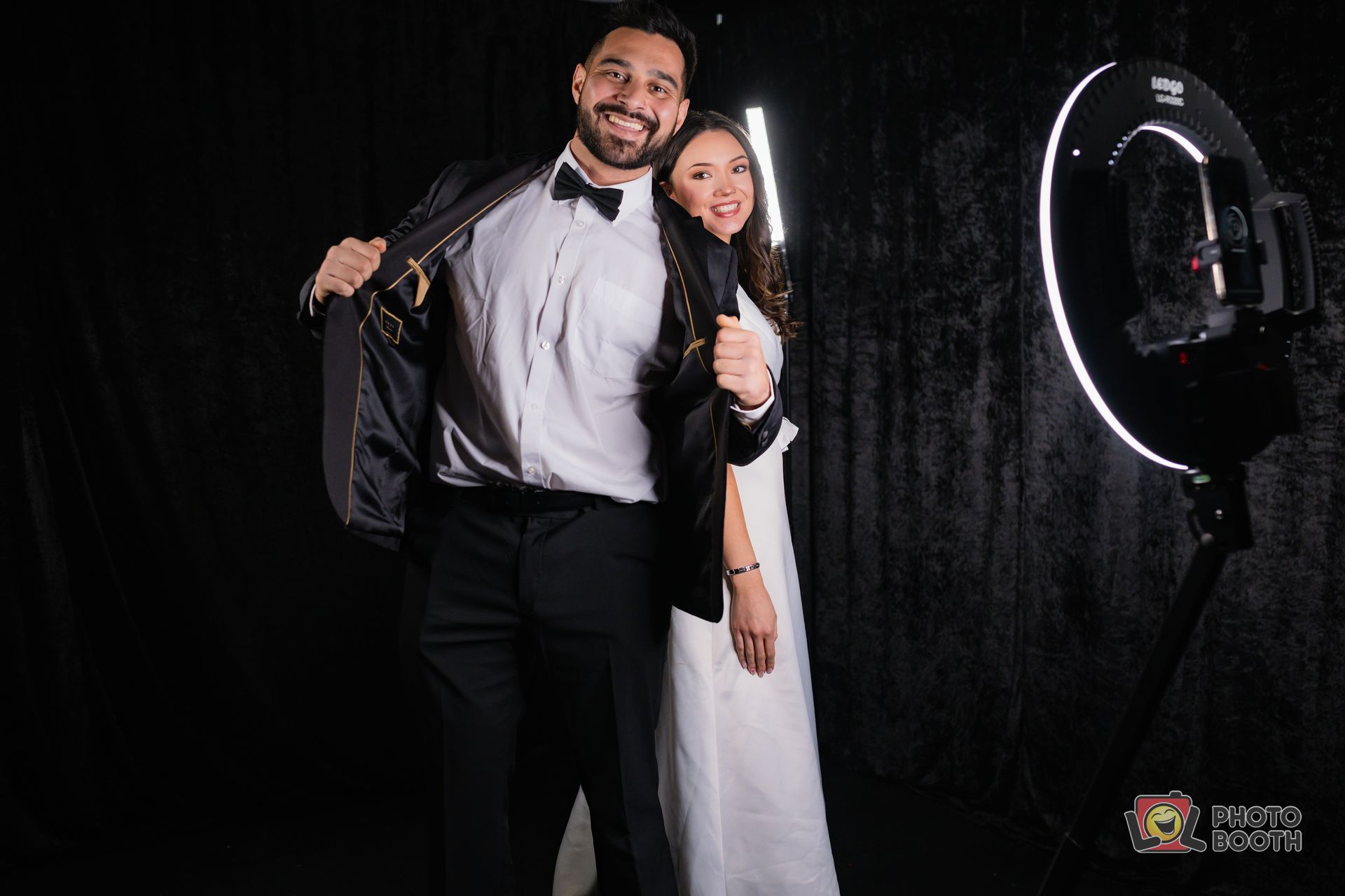 Couple in formal attire posing by a ring light. Man pulls back jacket. Black backdrop.