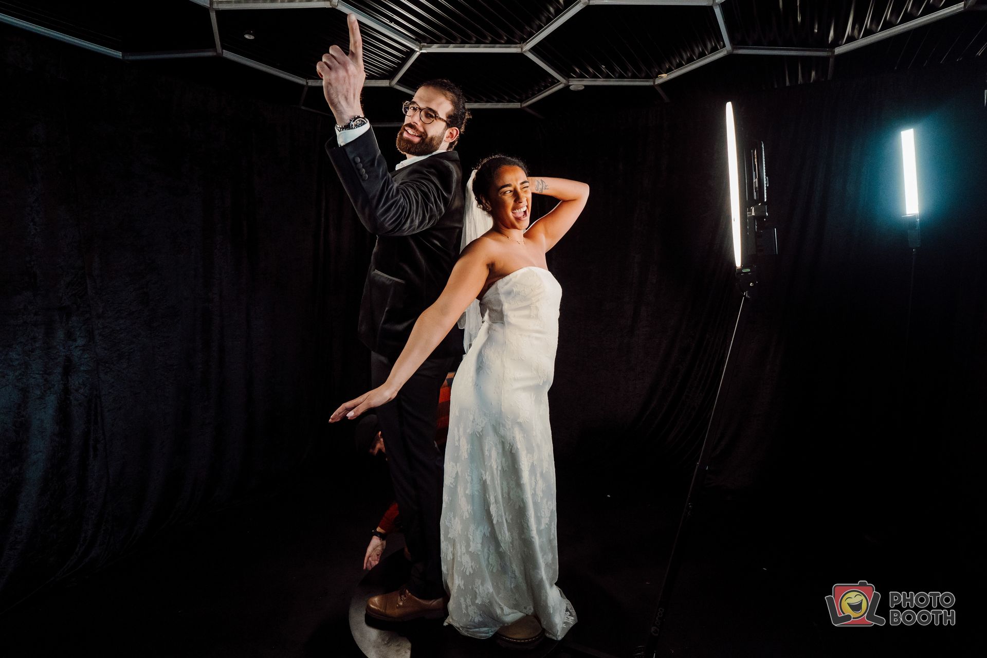 A couple, the bride in a white gown and groom in a suit, pose playfully in a black setting.
