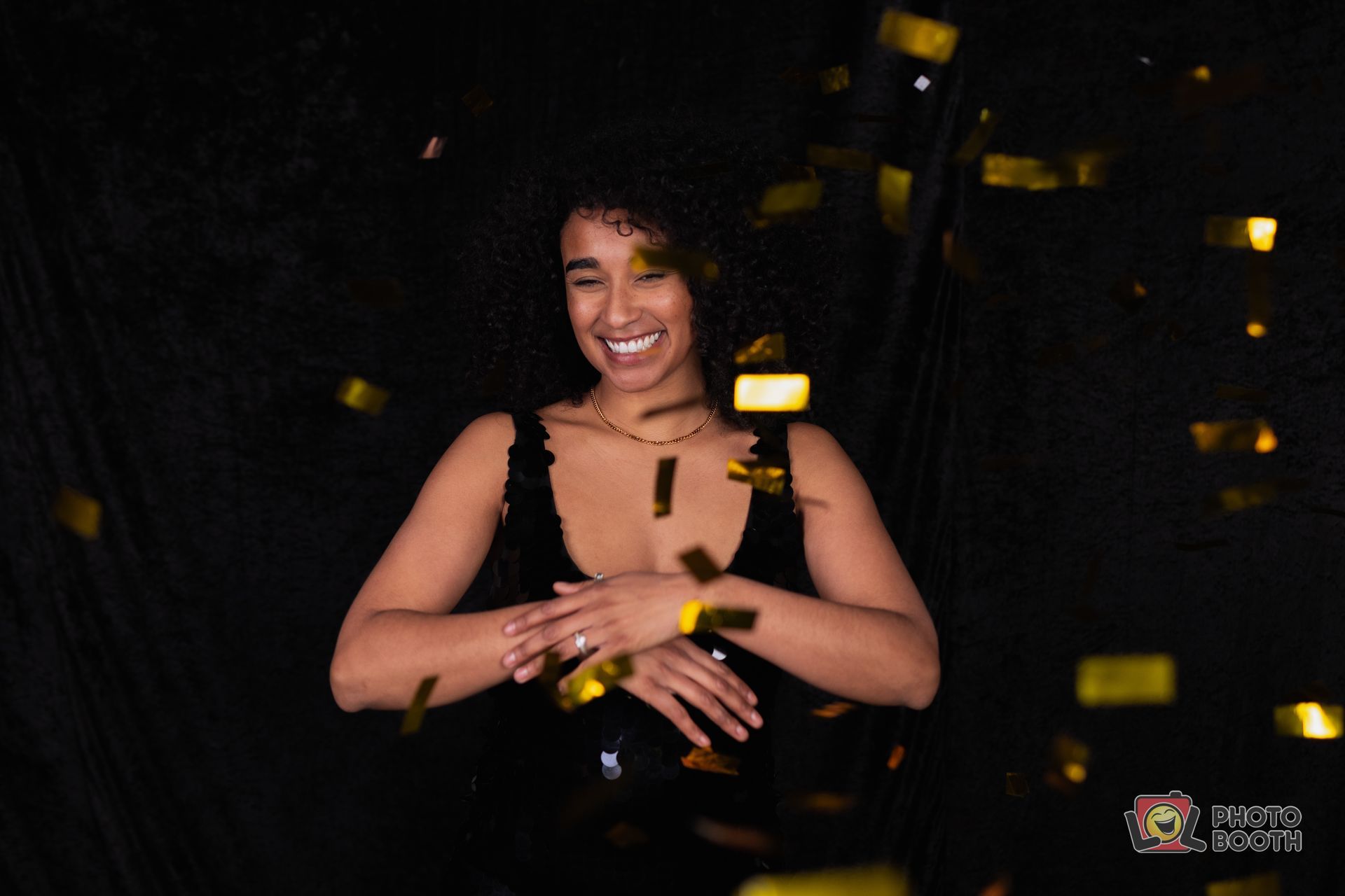 Woman in black dress smiling, surrounded by falling gold confetti, against black backdrop.