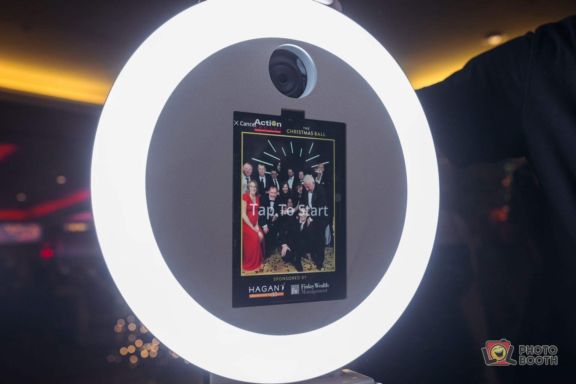 Photo booth with a bright ring light displaying a printed photo. A person's arm is visible.