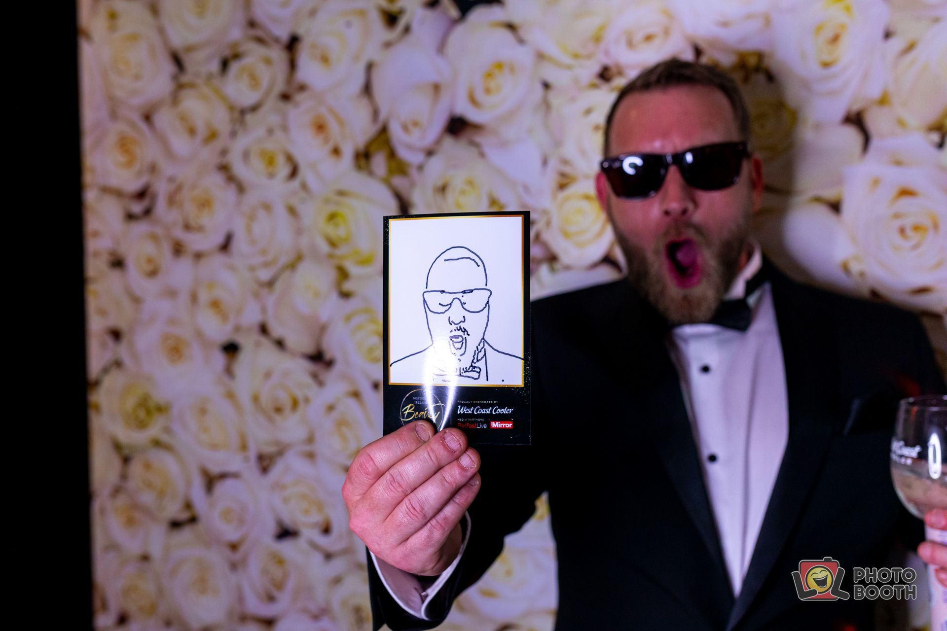 Man in tuxedo and sunglasses holds a drawing of himself, with white roses backdrop, open-mouthed expression.