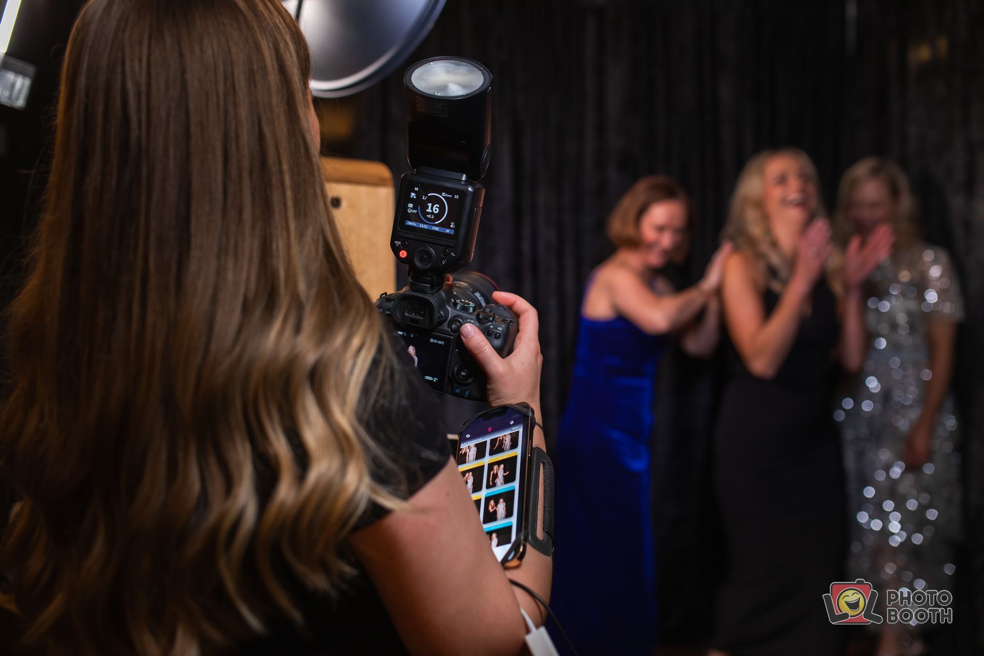 Photographer takes a photo of three women in formal attire; black backdrop.