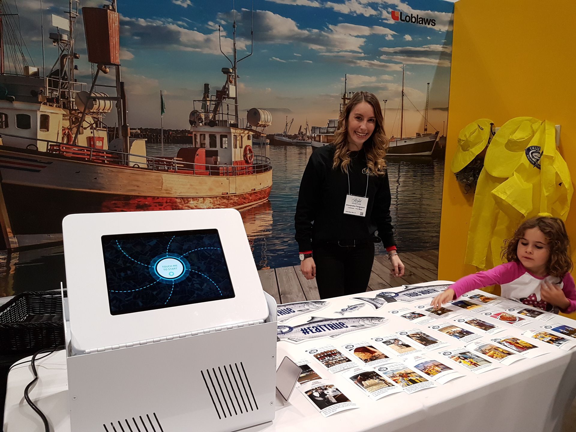 Woman and child at a booth with a touch screen display and printed materials, harbor background.