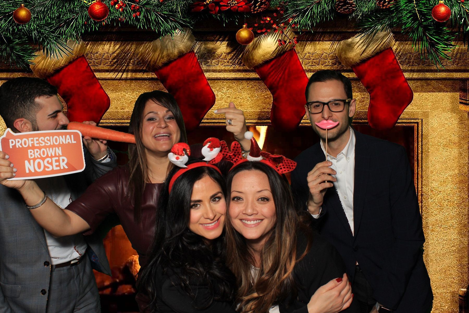 Group of people posing with props in front of a Christmas backdrop; holding a carrot nose, and lips.