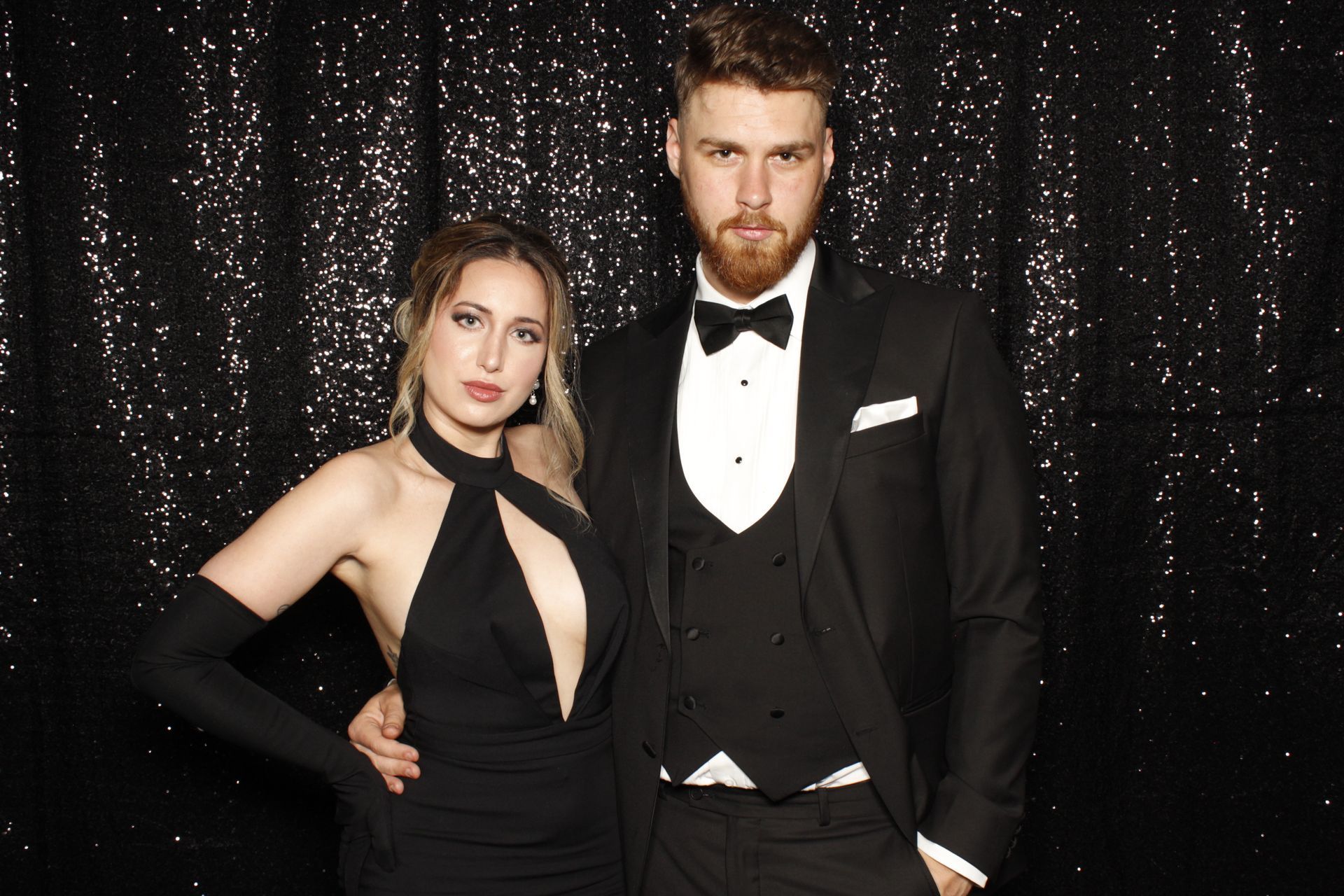 Woman in black gown and man in tuxedo pose in front of a sequined backdrop.
