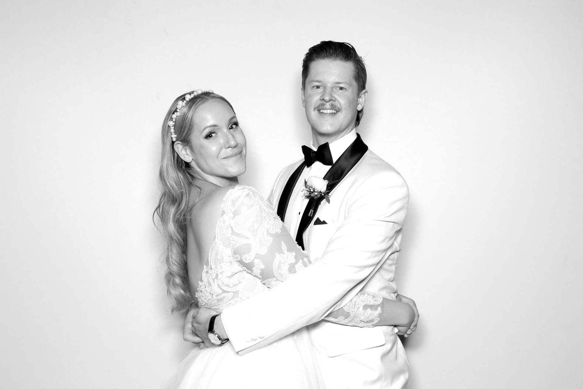 Bride and groom embrace, smiling. Bride wears a dress and headpiece, groom in a tuxedo. Black and white photo.