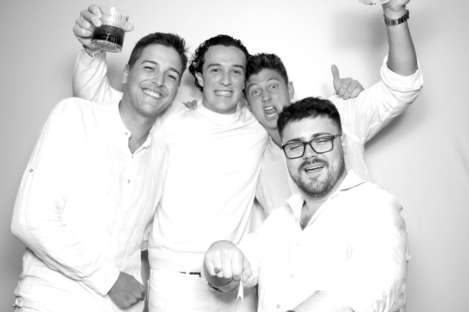Four men in white clothing, smiling and celebrating. One man holds a drink up, others make gestures.