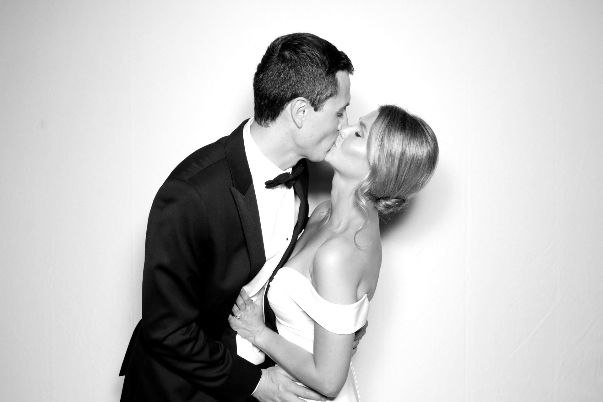 Couple kissing, man in tuxedo, woman in white dress, against white backdrop.