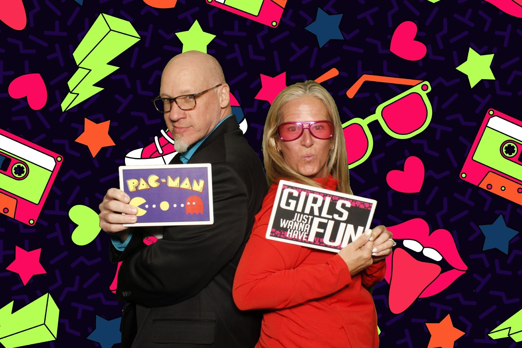 Man and woman pose with photo booth props against a retro background. Man holds Pac-Man sign, woman holds 