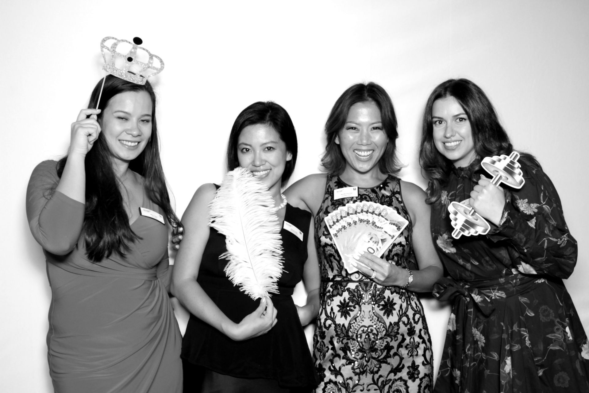 Four women pose for a photo with props in a photo booth, smiling against a white backdrop.