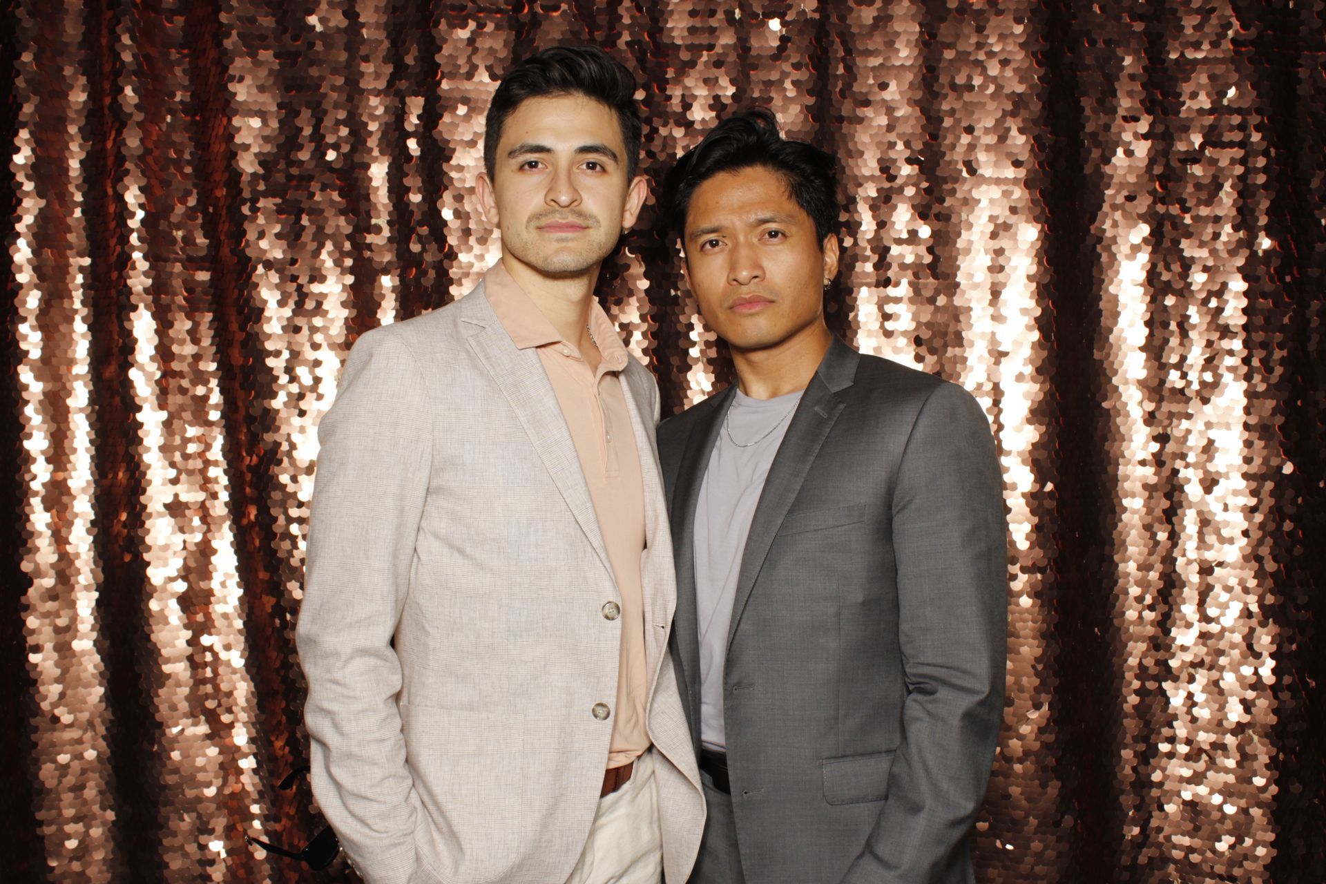 Two men posing in front of a sequined backdrop. One in light suit, the other in a gray blazer.