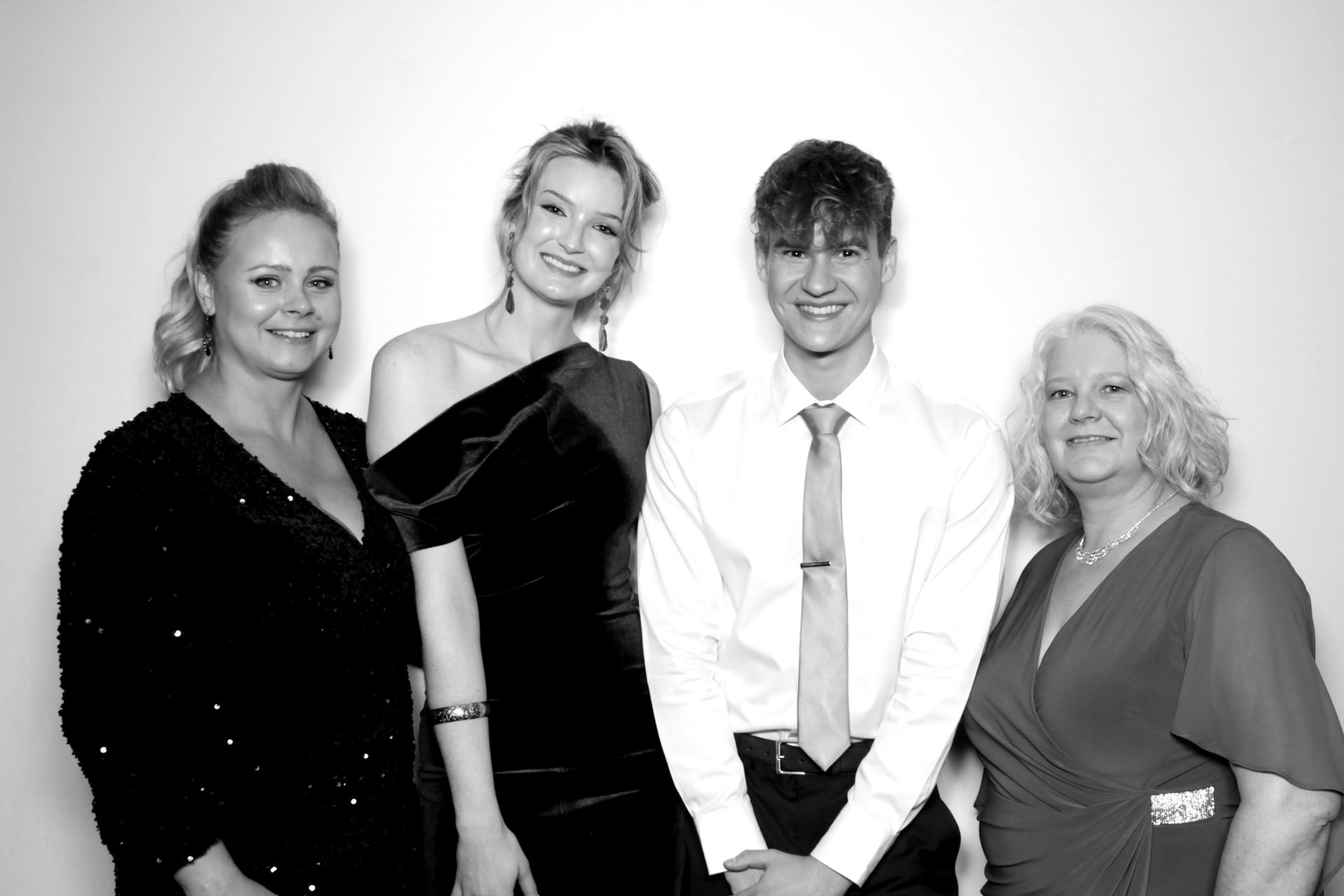 Four people smiling at a camera against a white backdrop. Woman in black gown, other in red. Man in white shirt and tie.