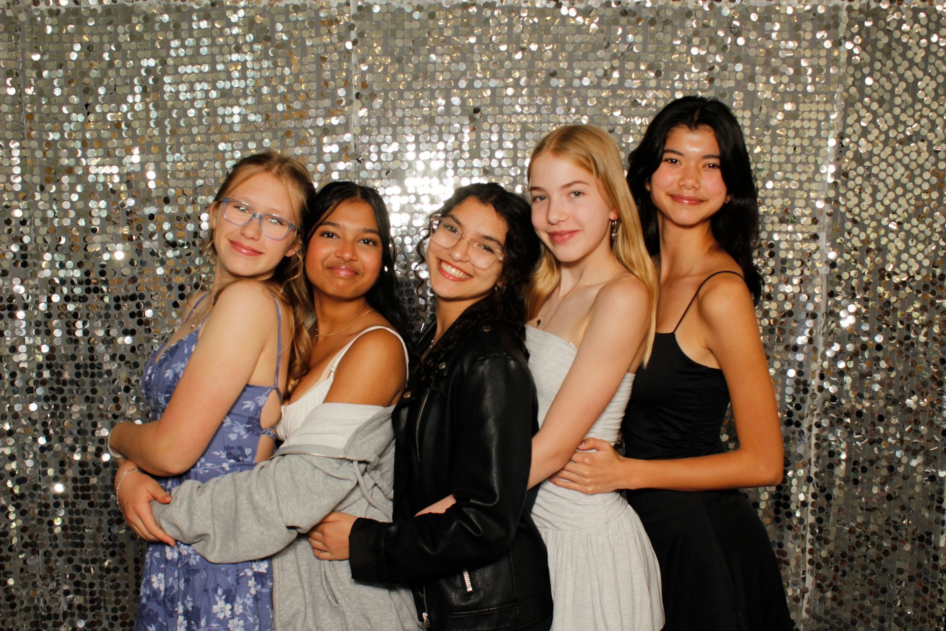 Five young women in formal attire pose in front of a silver backdrop. They smile and embrace each other.