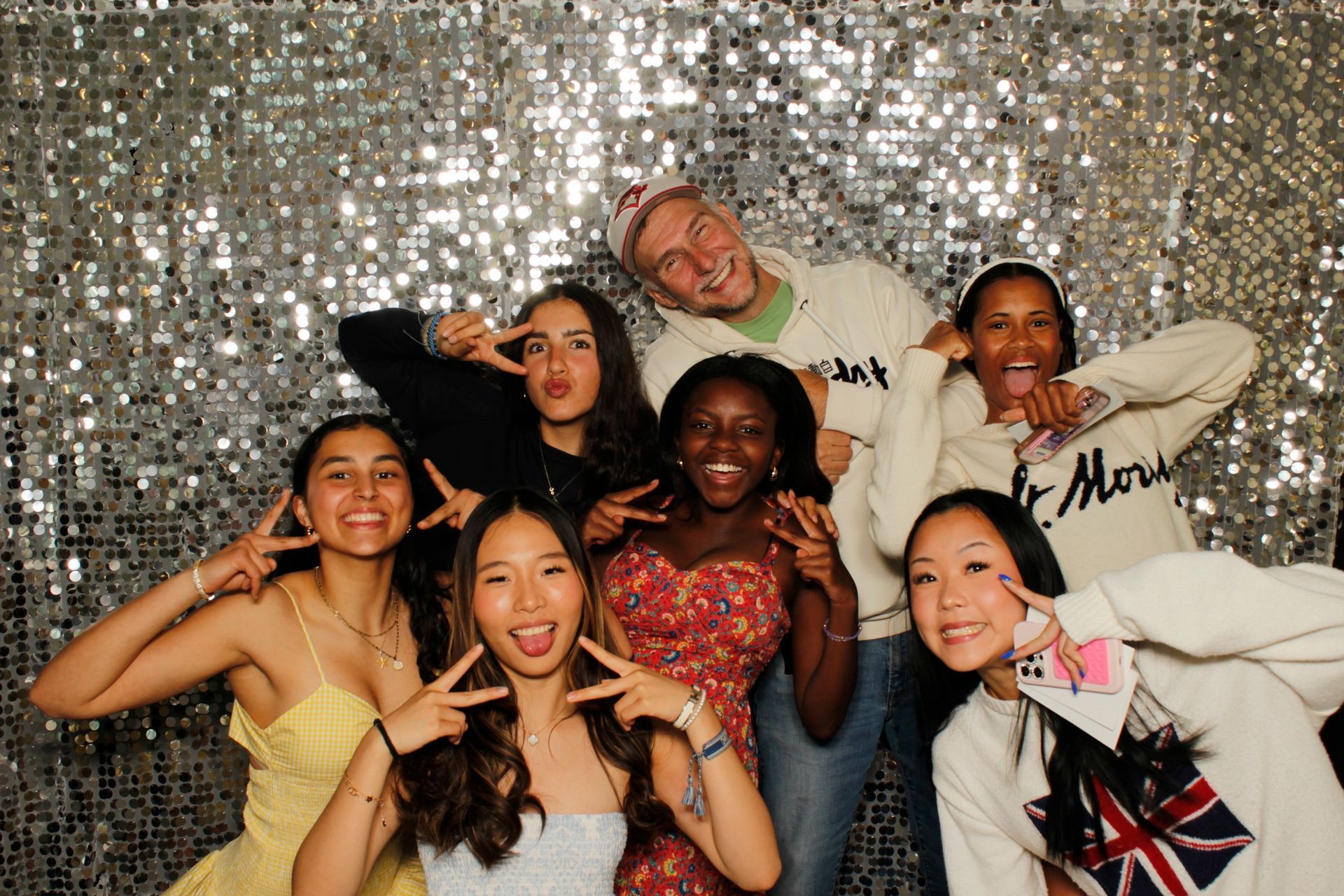 Group of friends posing for a photo. They smile, make peace signs, and stick out their tongues in front of a sequined backdrop.