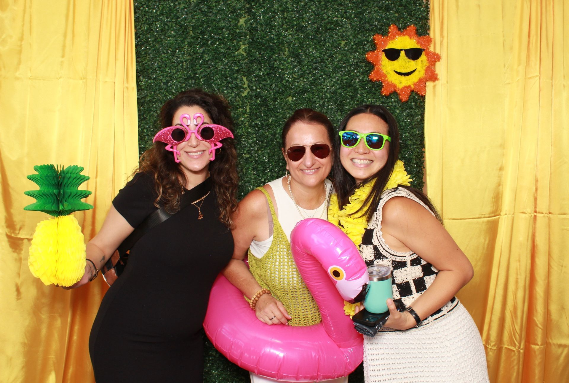 Three women pose in a photo booth with props. They are smiling and wearing sunglasses. A green wall is the backdrop.