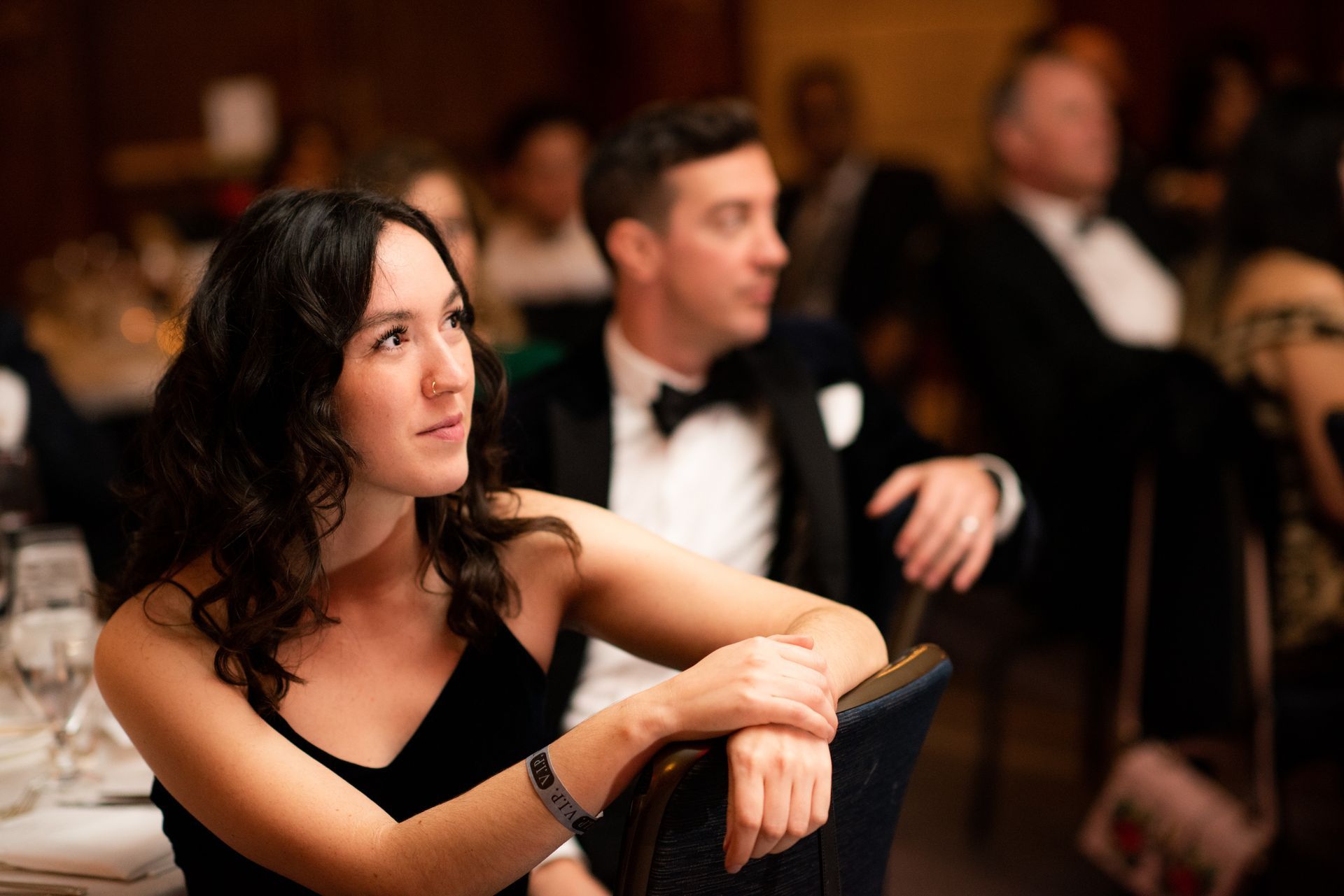 Woman in black dress at formal event, looking up, with people seated nearby.