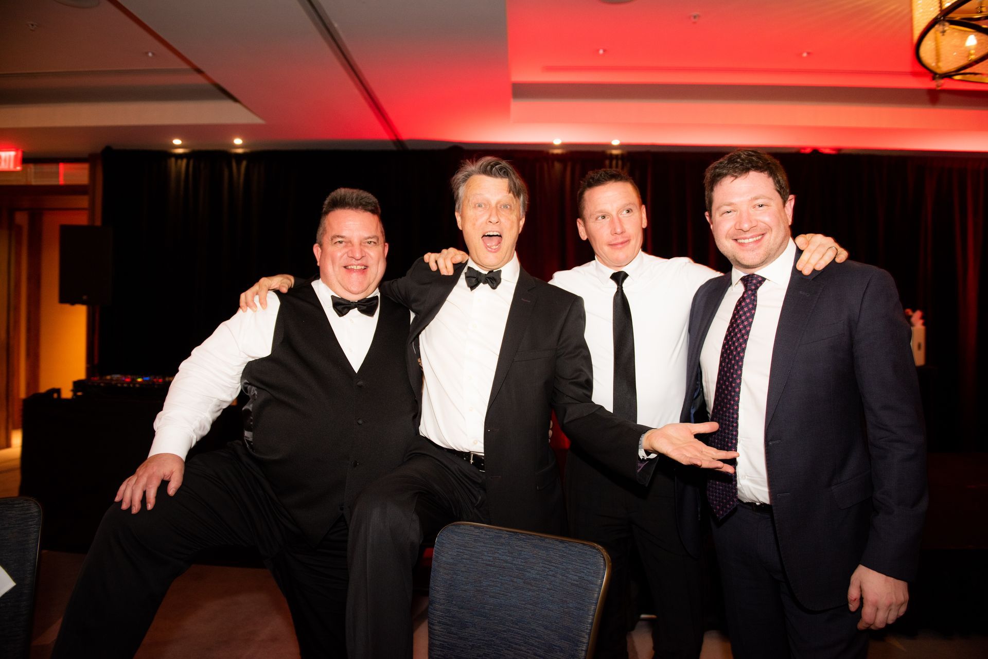 Four men pose for a photo at a formal event, smiling. One man squats, others stand, all in suits.