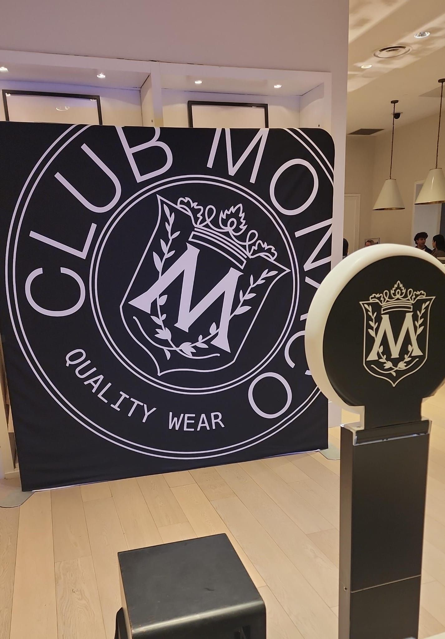 Club Monaco logo on a large black backdrop inside a store, with a decorative display stand in the foreground.
