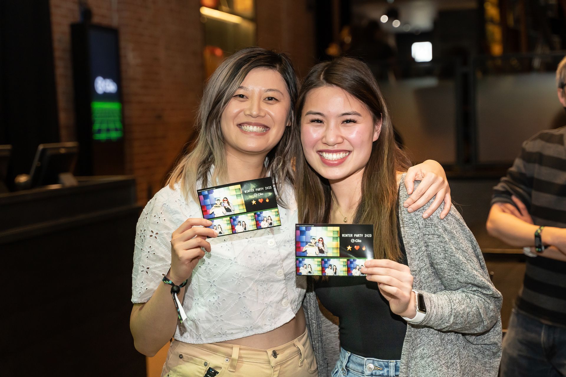 Two women smiling, holding photo strips. One with light hair, white top, khaki pants. The other with dark hair, black top, gray sweater.
