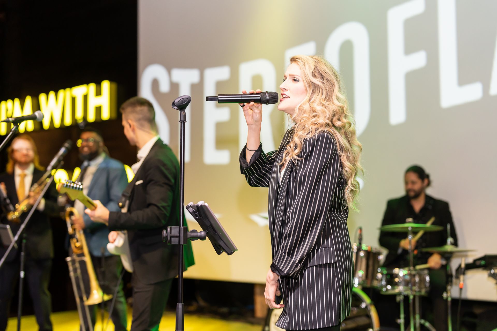 Band Stereo Floss performs live on stage. Singer in black and silver outfit, band in suits. Yellow background.