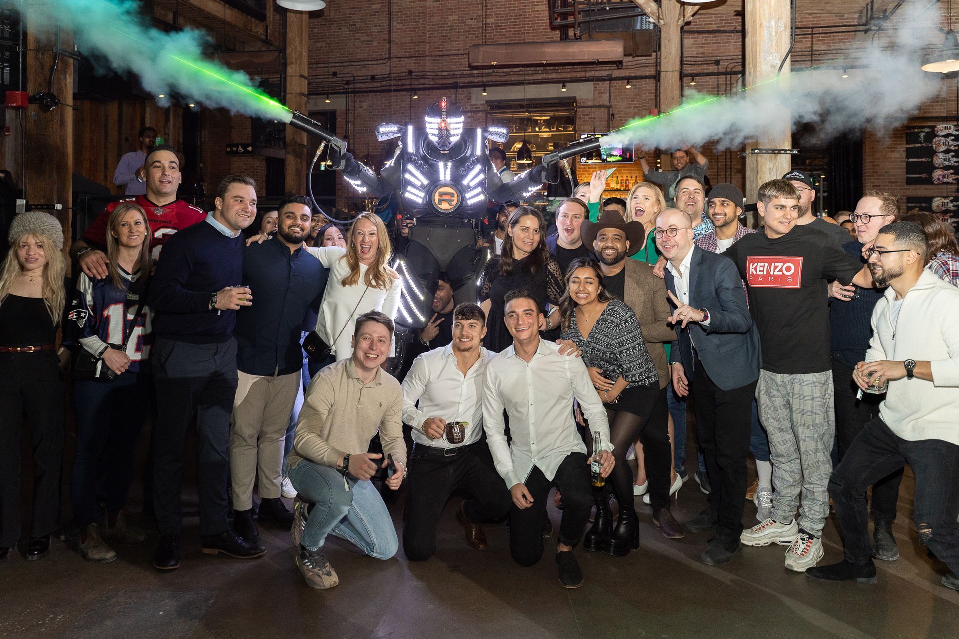 Group poses with a robot, green smoke, brick building background.